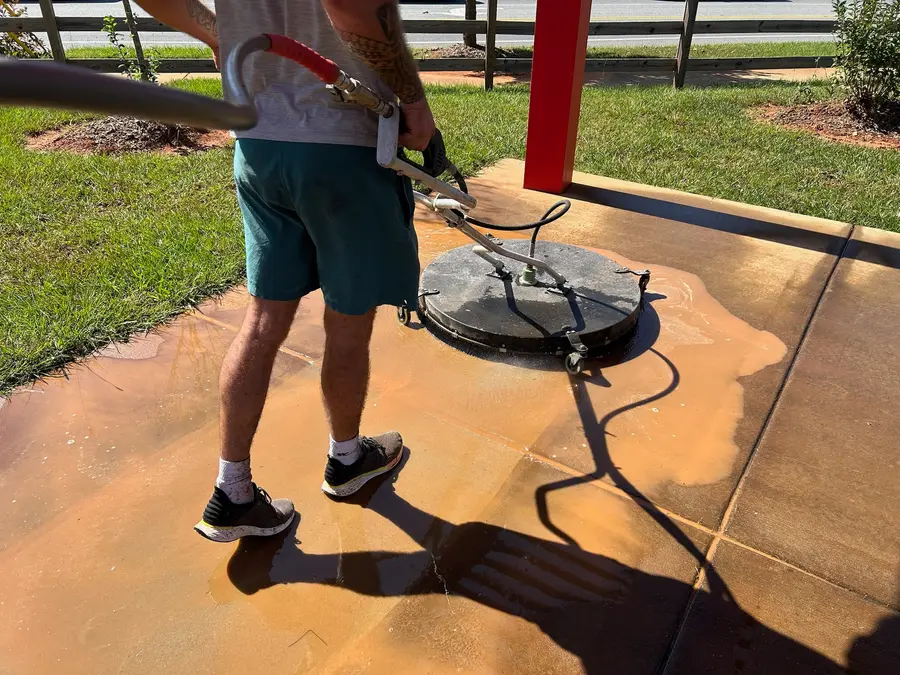Worker using a rotary surface cleaner on a concrete pad, rust-colored soap runoff visible.